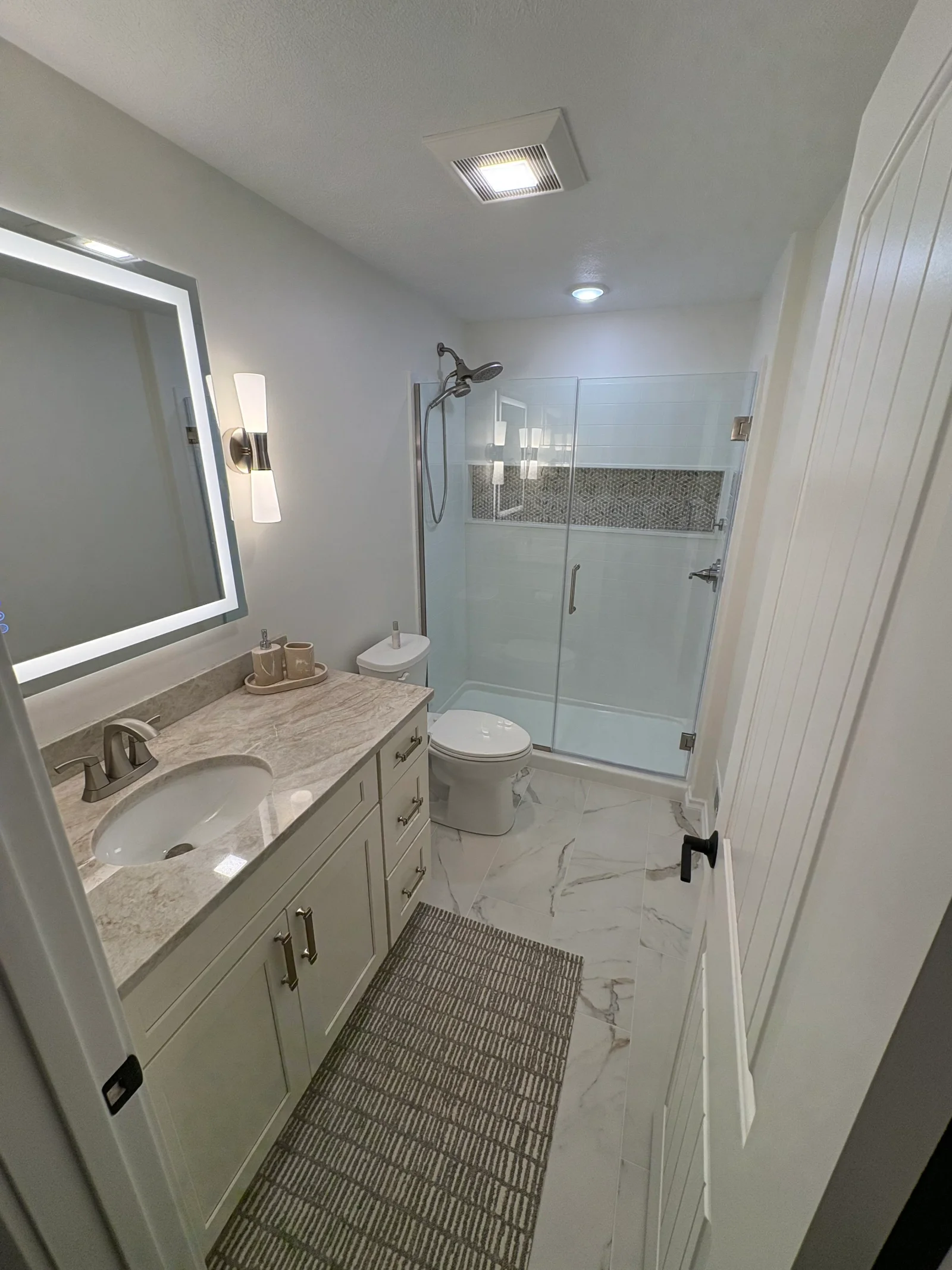 Remodeled bathroom with frameless glass shower, mosaic accent niche, marble-look floor tile, white shaker vanity, and LED mirror