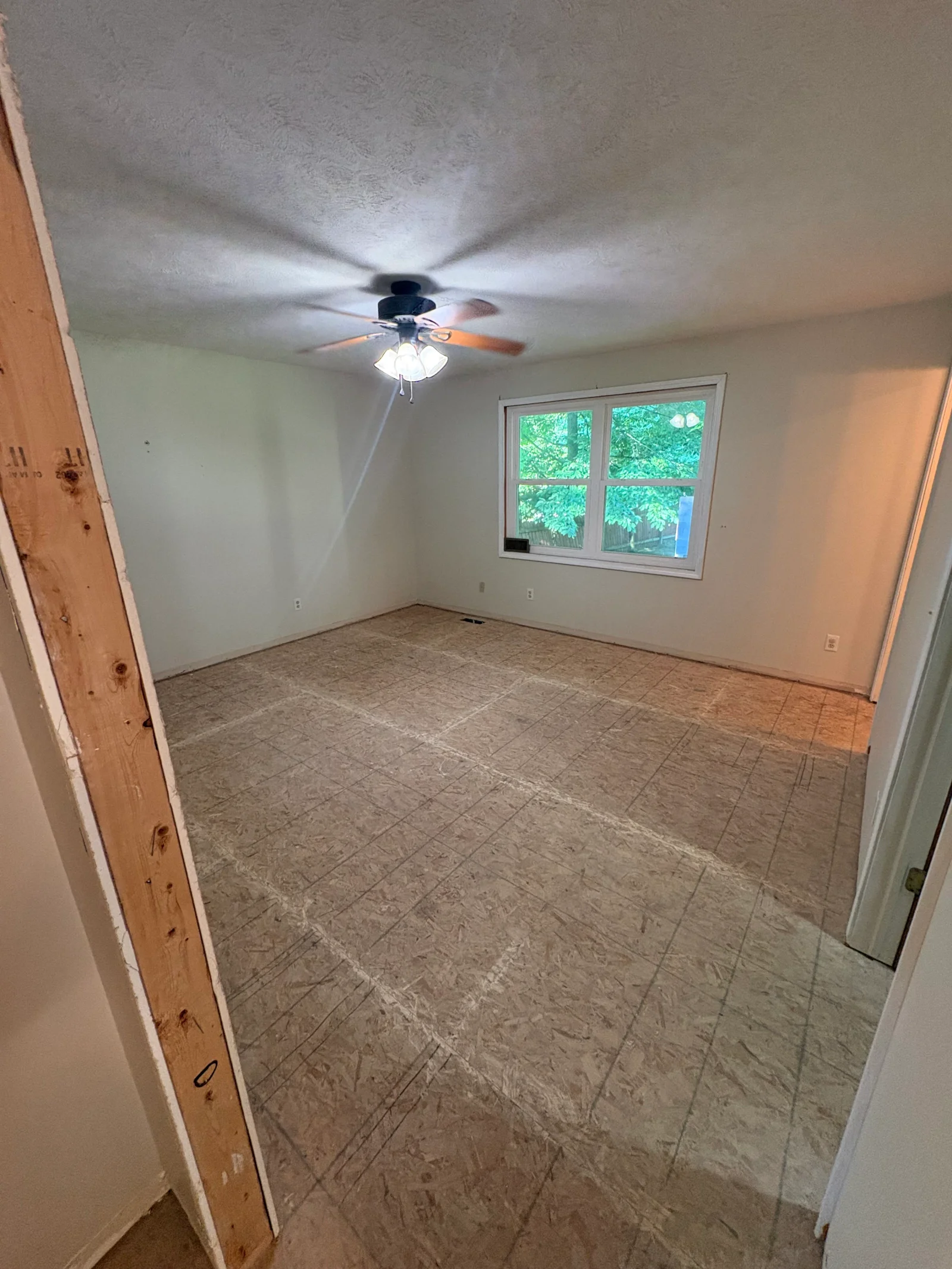 Empty bedroom with OSB subfloor layout lines, textured ceiling, ceiling fan, and white vinyl double-hung window