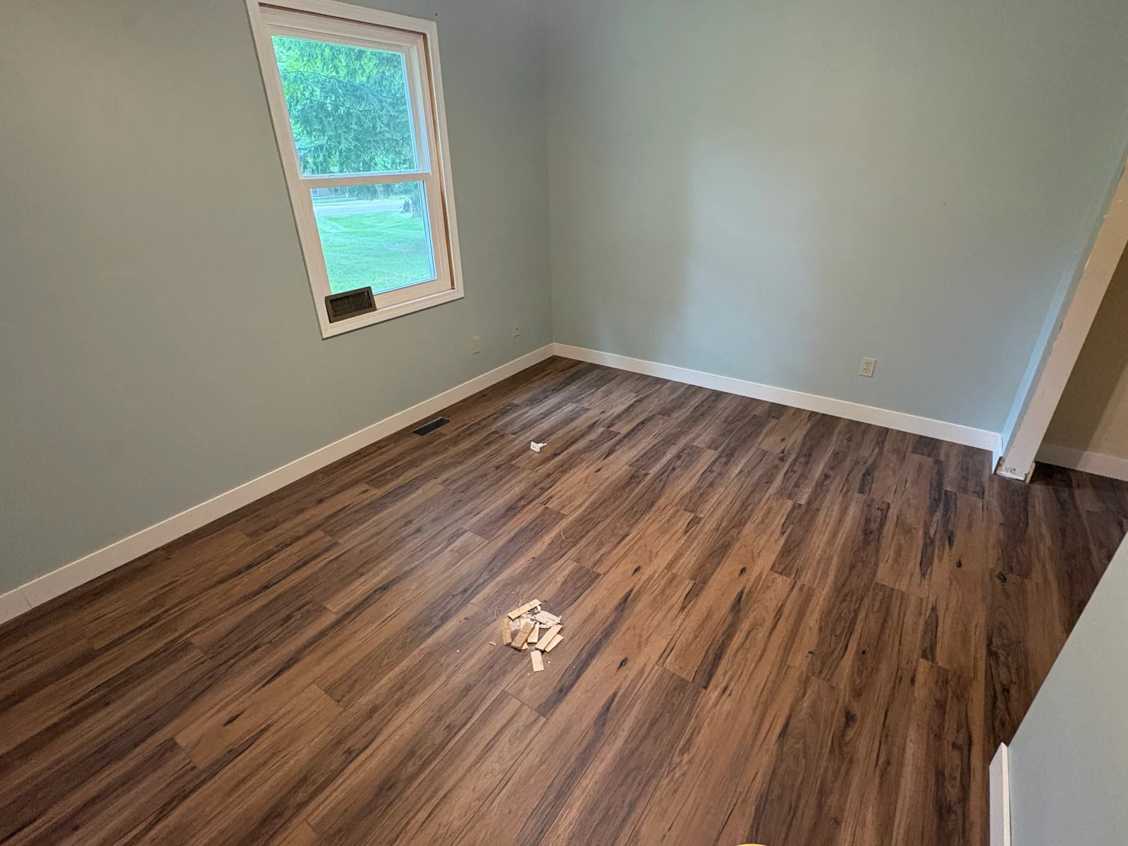Newly installed wood-look vinyl plank flooring in a bedroom with sage green walls, white baseboards, and a window with white trim