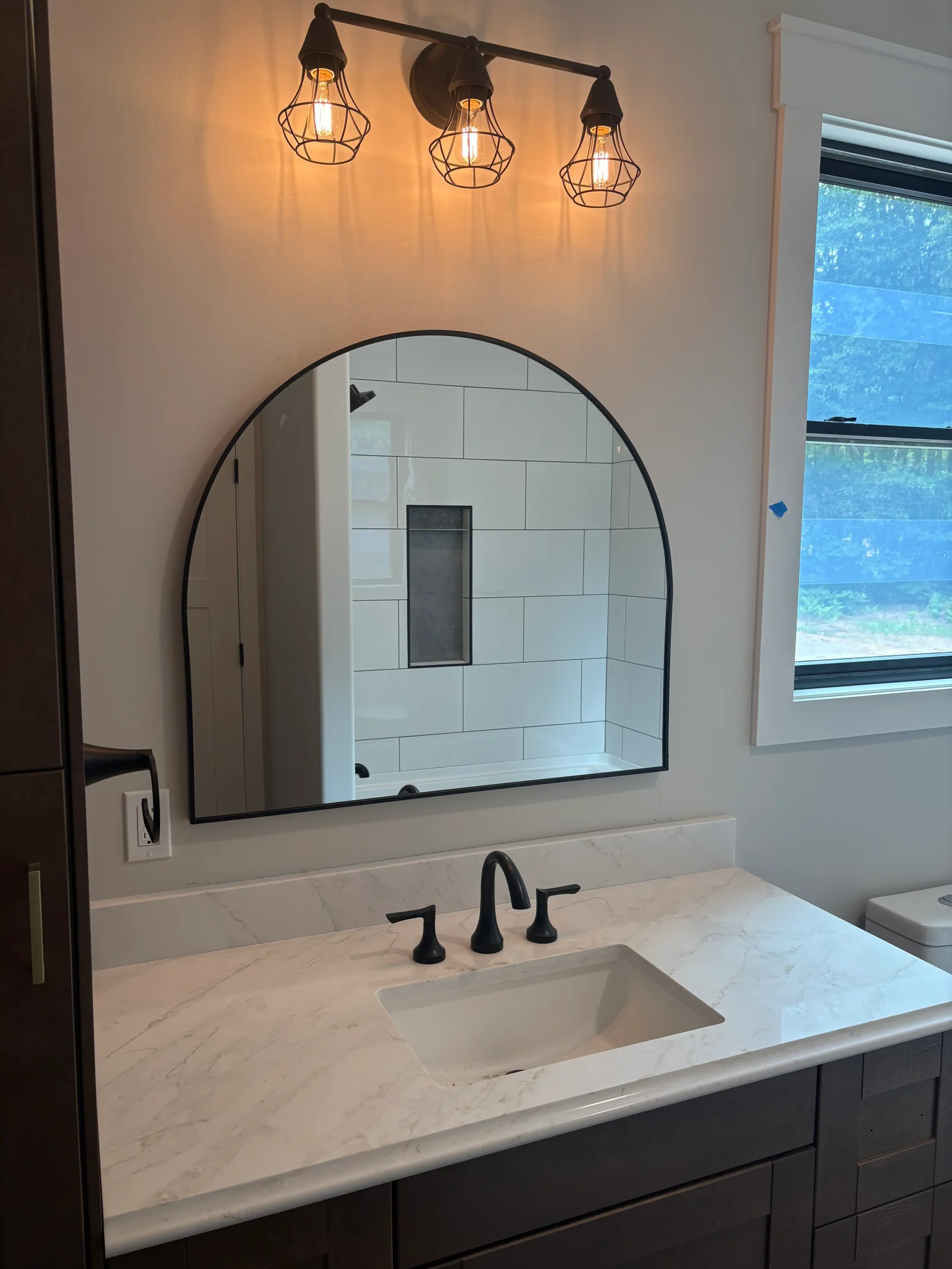 Bathroom vanity with white veined quartz countertop, undermount sink, matte black faucet, arched black-framed mirror, and caged Edison sconce lighting