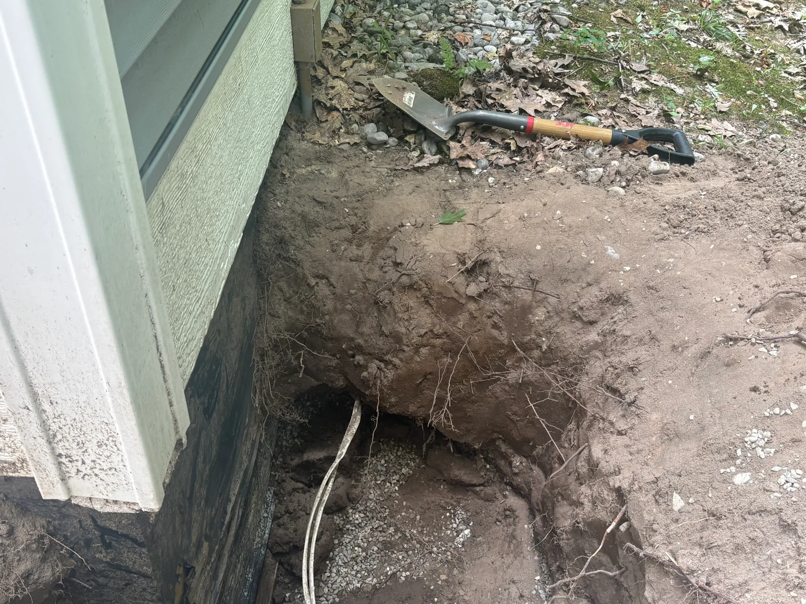 Excavated trench exposing dark foundation wall and gray conduit beside green siding, with gravel and shovel