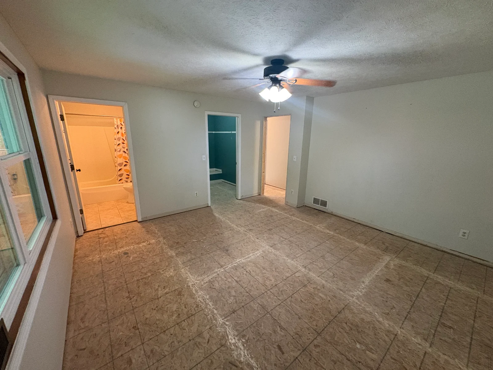 Empty bedroom with particleboard subfloor marked with chalk layout lines, ceiling fan, and view into attached bathroom with tub-shower