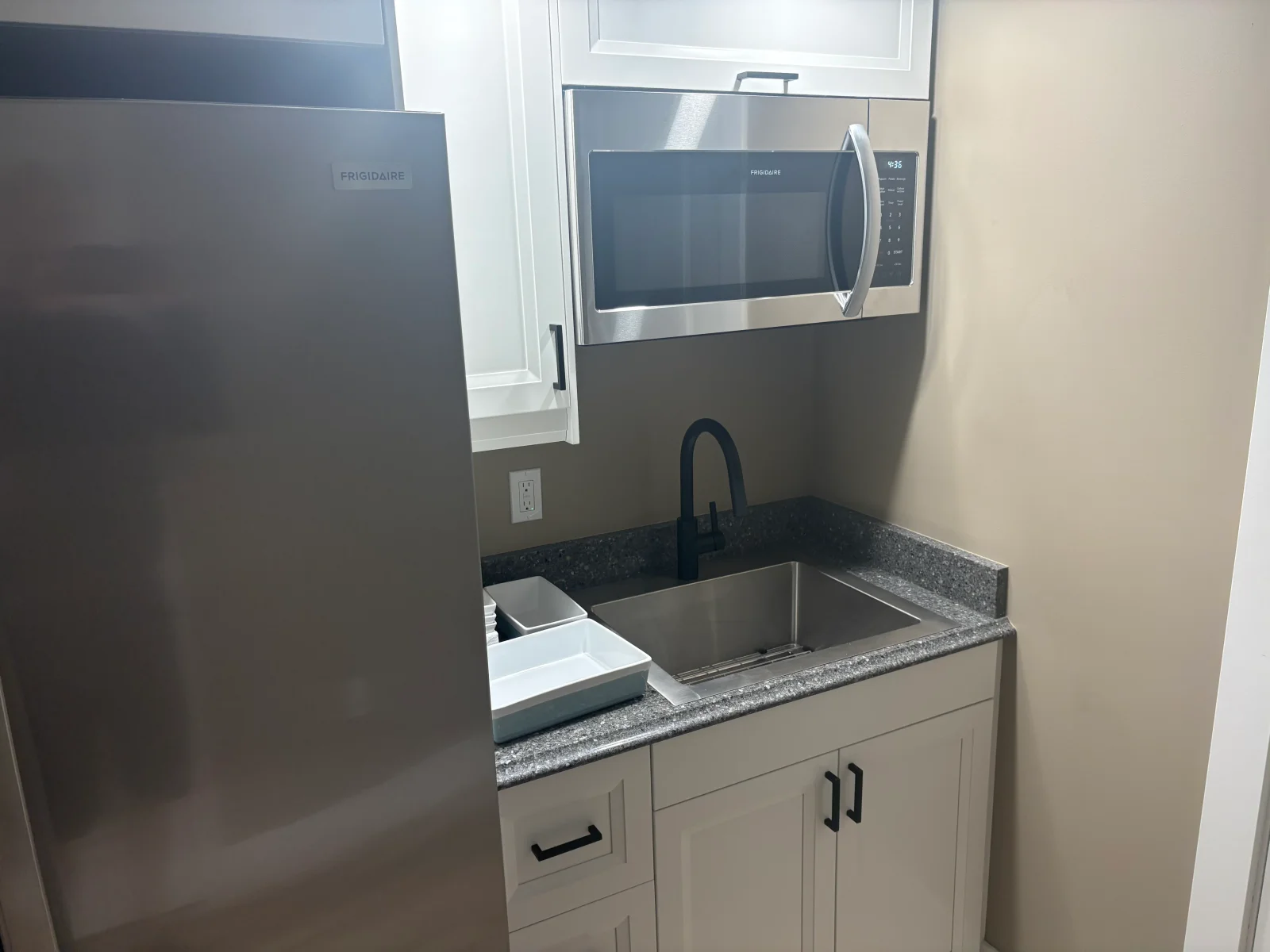 Compact kitchenette with white shaker cabinets, black hardware, speckled granite countertop, stainless undermount sink, and stainless microwave