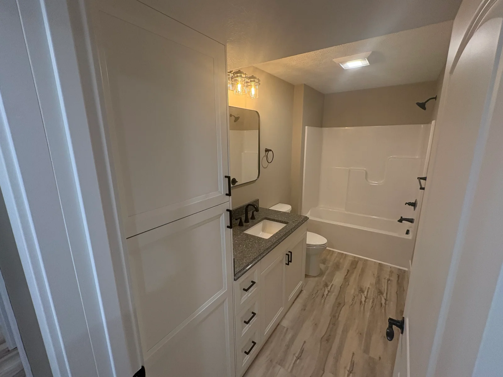 Bathroom with white shaker vanity, speckled granite countertop, black fixtures, tub-shower combo, and wood-look vinyl plank flooring