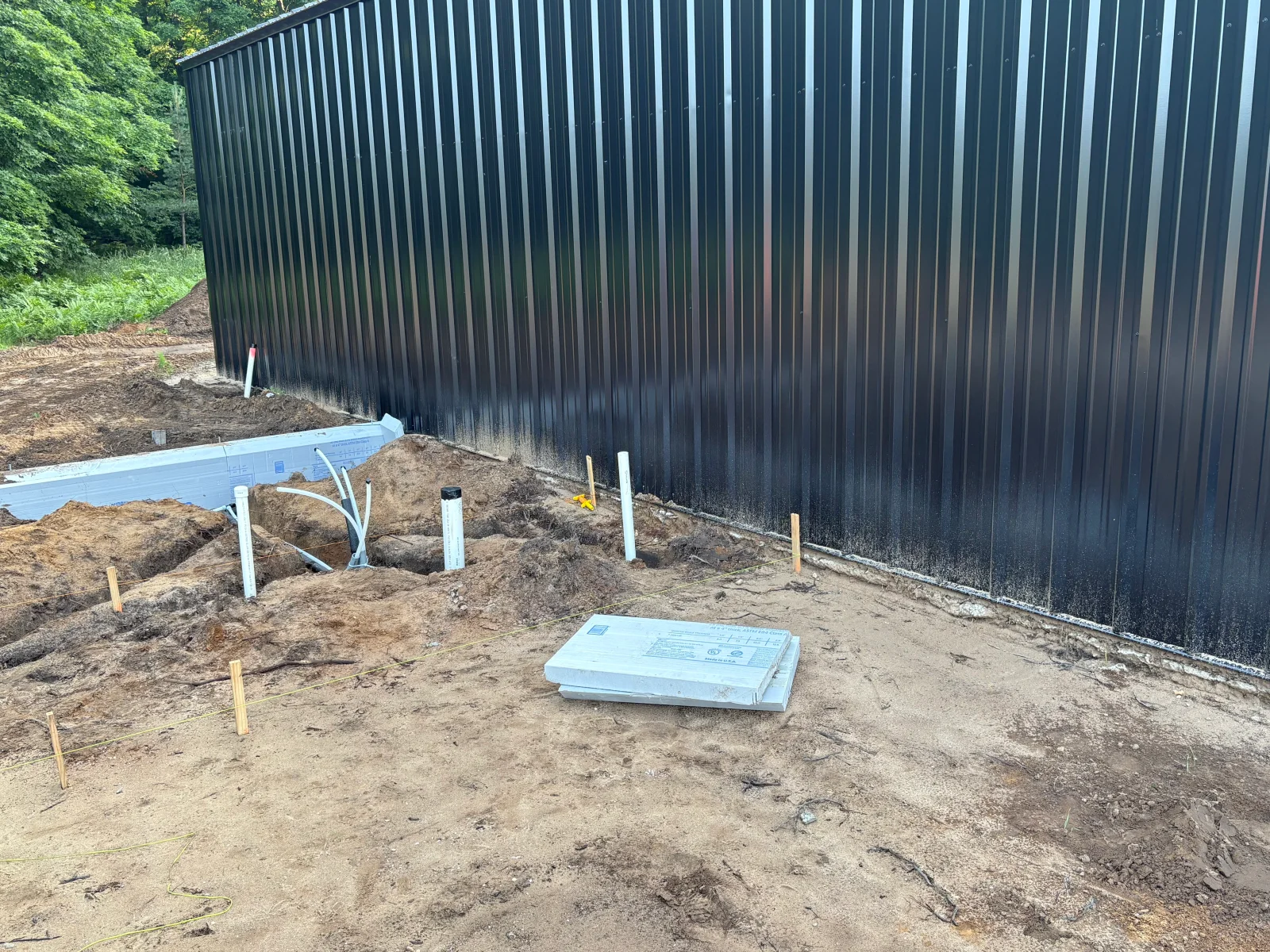 Excavated trench beside dark corrugated metal building with PVC pipe, rigid foam insulation panels, and staked layout lines