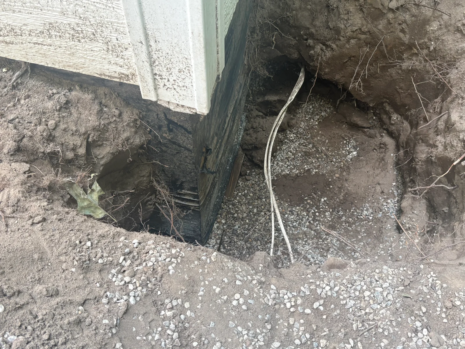 Excavated corner of wood sill and vinyl siding with exposed gravel and metal cables at grade