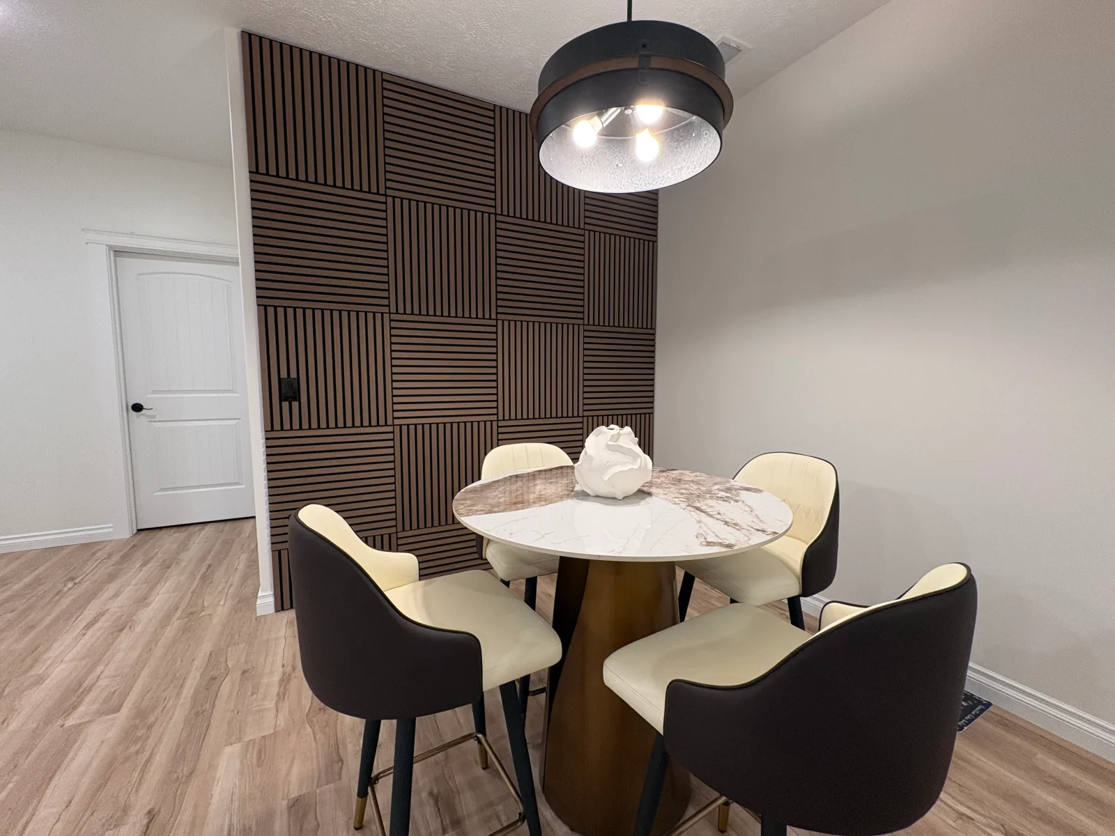Dining nook with dark slatted wood accent wall, marble-top pedestal table, cream and charcoal bar stools, and vinyl plank floors