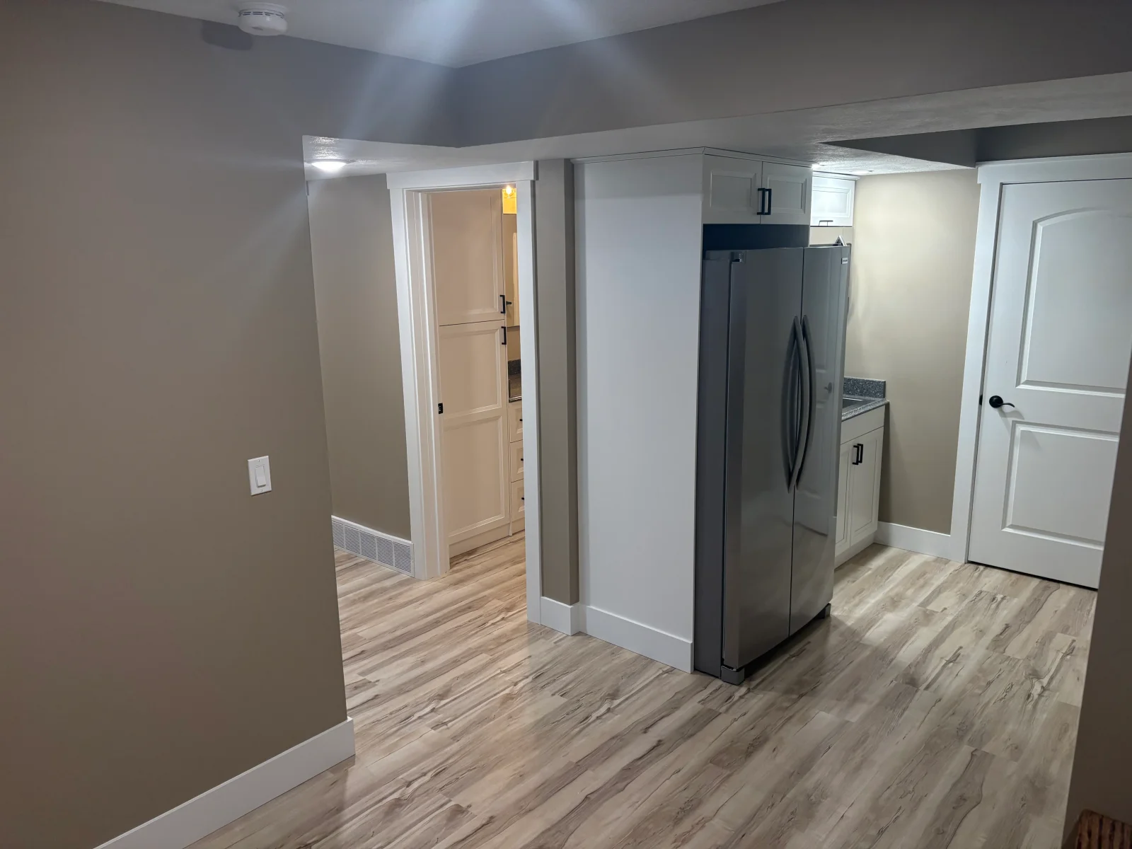 Basement kitchenette with white shaker cabinets, stainless refrigerator, gray countertop, and wood-look vinyl plank flooring