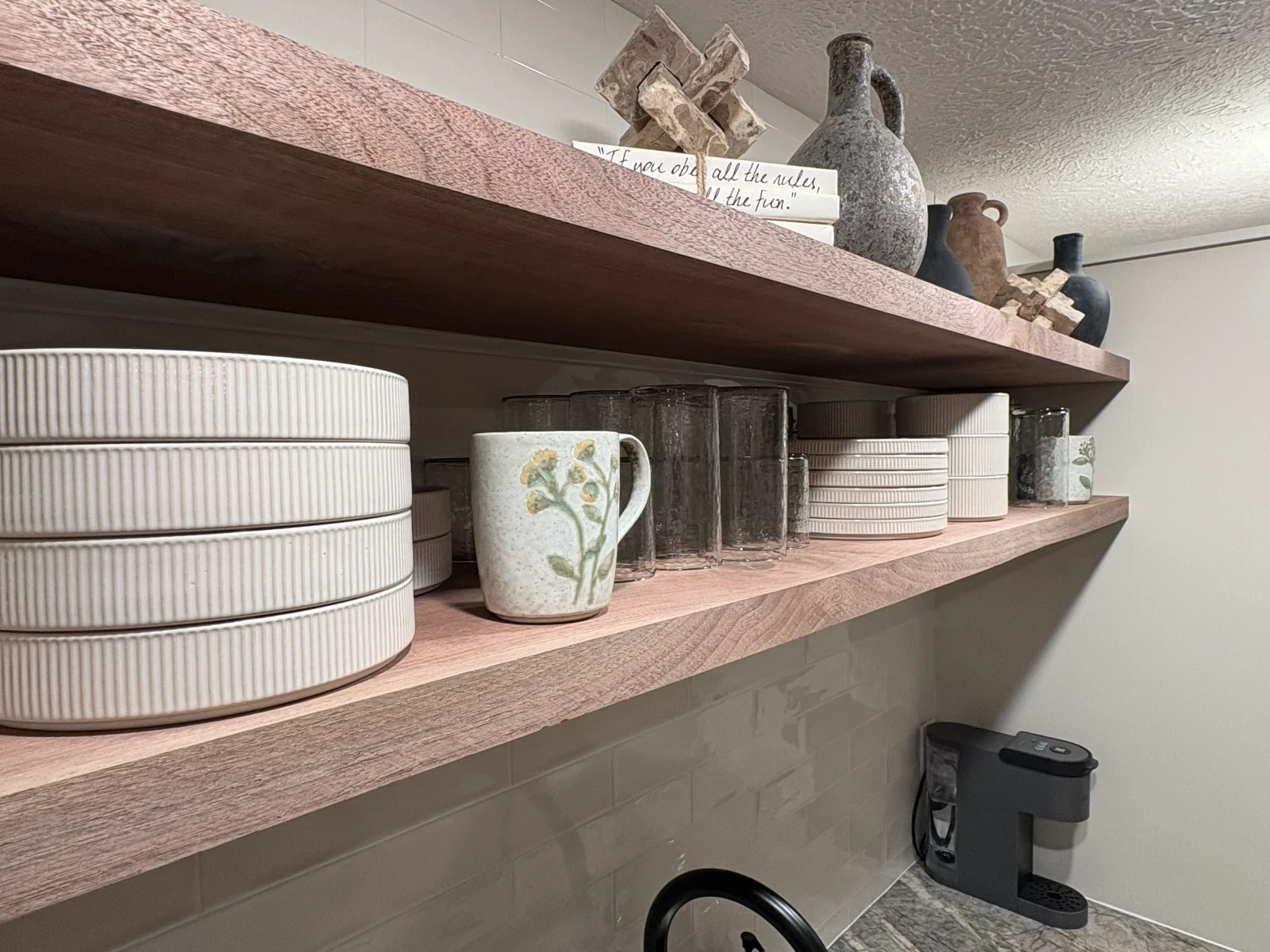Solid wood floating shelves with ceramic dishware, glassware, and decorative stoneware above gray subway tile backsplash