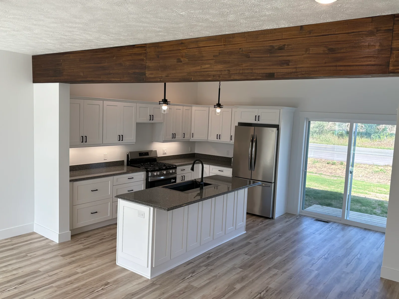 White shaker kitchen with dark quartz island, black pendant lights, stainless appliances, and wood-look vinyl plank flooring
