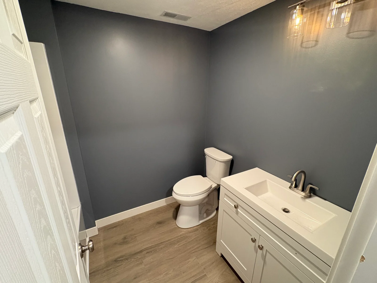 Powder room with wood-look vinyl plank flooring, slate-blue painted walls, white vanity with brushed nickel faucet, and white toilet