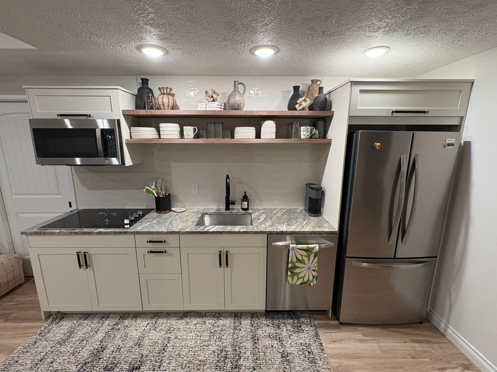 Cream shaker cabinets, speckled stone countertop, black induction cooktop, stainless appliances, and walnut floating shelves in a compact kitchenette
